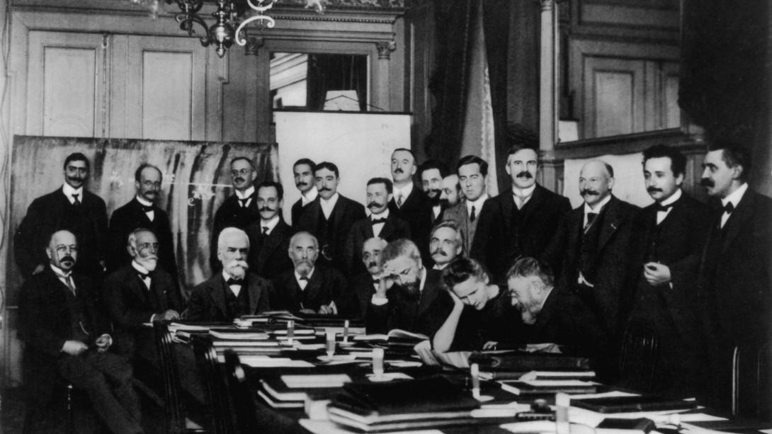 Participantes del I Congreso Solvay (1911) sobre «la radiación y los cuantos». Curie (sentada, segunda a la derecha y con la cabeza apoyada sobre su mano) conversa con Henri Poincaré; detrás de ellos, un joven Albert Einstein